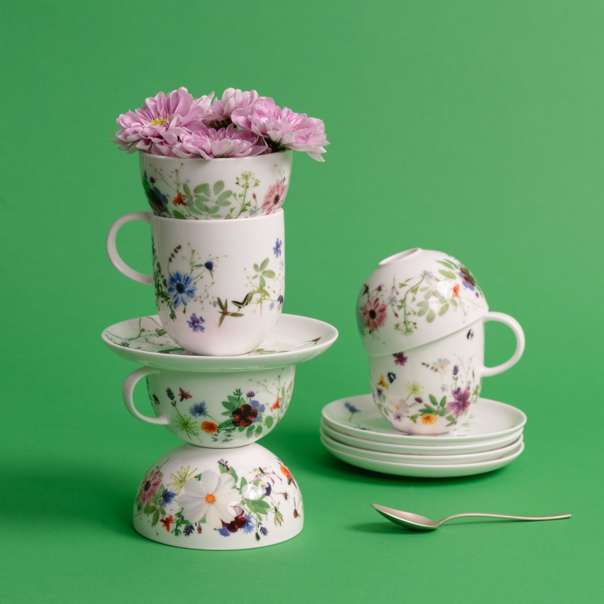 Stack of mugs, cups and small bowls from the floral Brillance Grand Air collection against a green background.